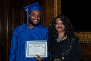 Two people standing and smiling at the camera. One person holding a diploma.