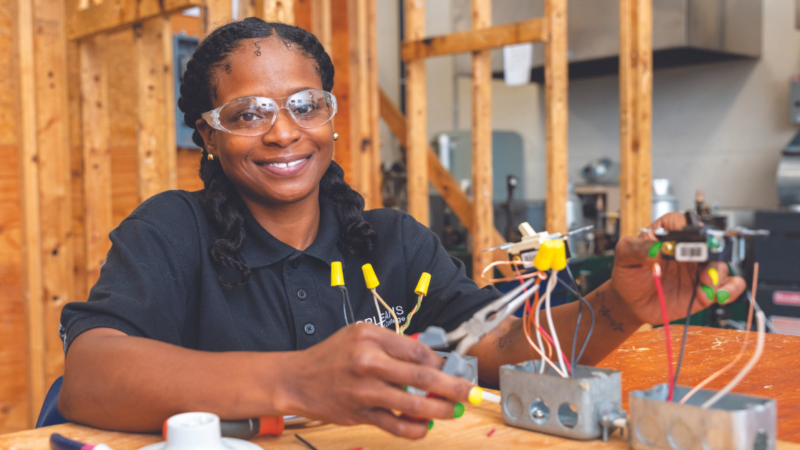 Image description. A woman in one of the programs at JEVS sits at a table working with wires. Her hair is in two braids, and she is wearing a black shirt and protective goggles. End description.