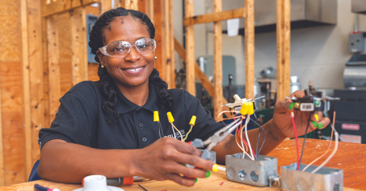 Image description. A woman in one of the programs at JEVS sits at a table working with wires. Her hair is in two braids, and she is wearing a black shirt and protective goggles. End description.