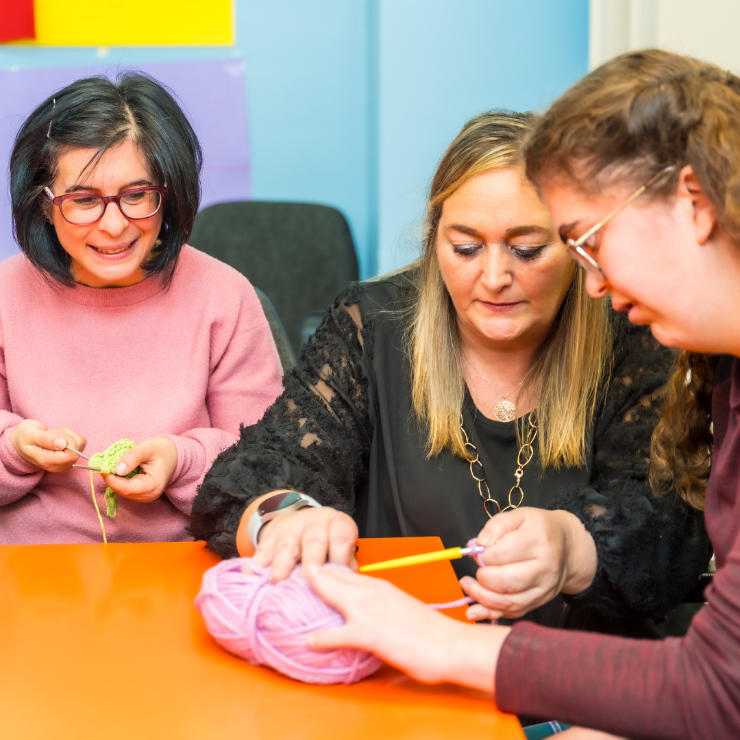 Image of three women sitting at a table; one woman is teaching the other two women how to crochet.