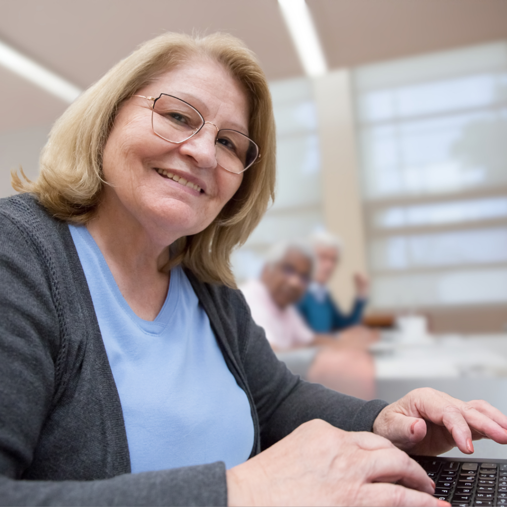 A woman sits at a desk typing while slightly turning to smile at the camera. Two people sitting in the background also turn toward the camera.