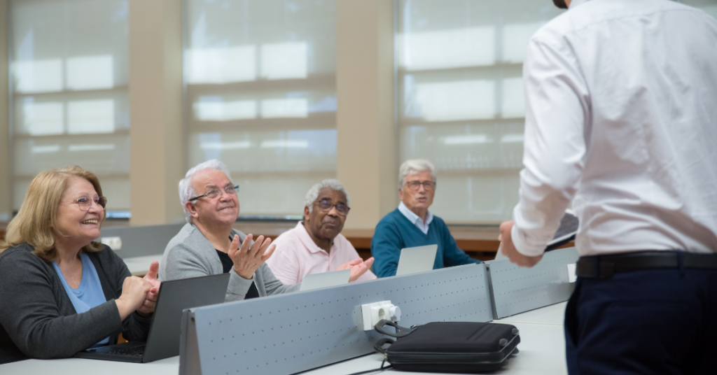 Four adults who are 55 and older sit at a table in a row while listening to a speaker who is standing in front of them.