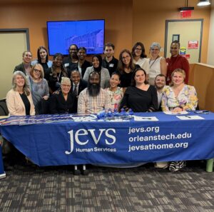 Members of the JEVS START Team and partners smile for the camera. They appear for a group photo behind a long table with a blue tablecloth that says JEVS Human Services and gives the URL for jevs.org; orleanstech.edu; jevsathome.org
