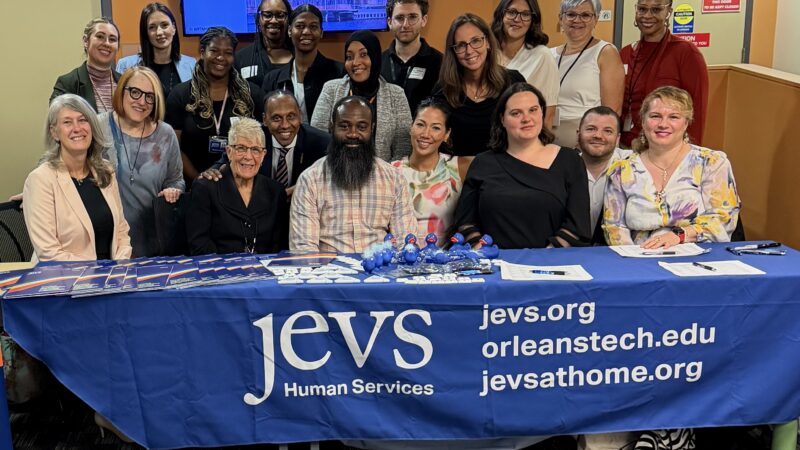 Members of the JEVS START Team and partners smile for the camera. They appear for a group photo behind a long table with a blue tablecloth that says JEVS Human Services and gives the URL for jevs.org; orleanstech.edu; jevsathome.org