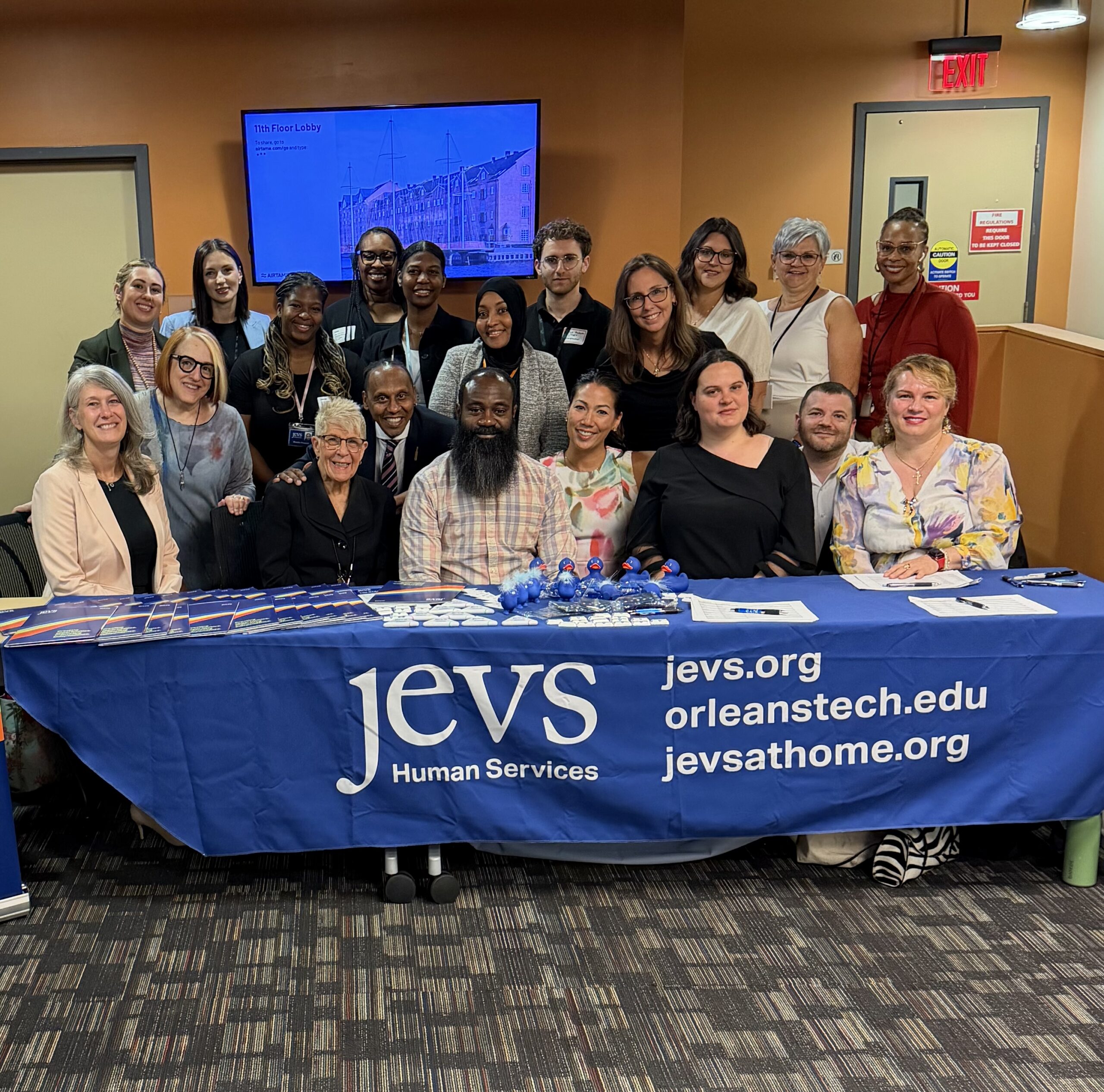 Members of the JEVS START Team and partners smile for the camera. They appear for a group photo behind a long table with a blue tablecloth that says JEVS Human Services and gives the URL for jevs.org; orleanstech.edu; jevsathome.org